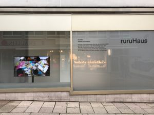 Video presentation by Gudskul in the ruruHaus window. In the video work, people can be seen bent over a table with printed materials. The entire shop window with the inscription ruruHaus slightly mirrors the stores opposite in Kassel's city center.