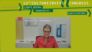 General Director of documenta and Museum Fridericianum gGmbH at the virtual 12th KulturInvest! congress. She wears glasses and short hair, a red jacket, in the background older documenta posters from past exhibitions.