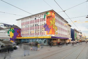 ruruHaus in Kassel: The building has many windows, left and right on the windowless areas the building is painted with the contrasting colors yellow, purple, green and orange. Illustrated with many hands grasping each other. In front of the building people are walking in the pedestrian zone.
