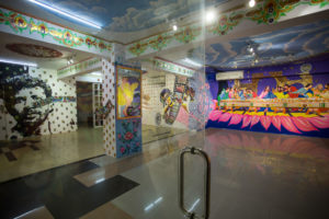The photo shows a colorful entrance hall of a cinema building, the glass door in the foreground. The walls are painted with various motifs from the art, pop and film scene, such as Marylin Monroe in a golden frame or the Last Supper motif; here with bright pink, blue, yellow colors on large wall.