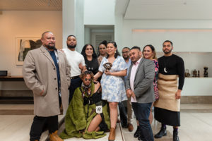 The group photo shows the artists' collective FAFSWAG. All look friendly, standing into the camera. One person squats in the middle, he/she has al single sunglasses on and has a striking green plush coat from which his/her thigh is visible. The woman next to it, standing in the middle, holds a prize in her hand and the other around the squatting person. All stand in a pleasantly bright room, in the background small sculptures and a modern drawing.