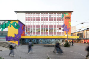 ruruHaus in Kassel: The building has many windows, left and right on the windowless areas the building is painted with the contrasting colors yellow, purple, green and orange. Illustrated with many hands grasping each other. In the center is a longer yellow bar that indicates the times of the documenta runtime. In front of the building people are walking in the pedestrian zone.