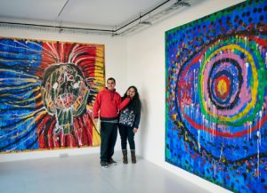 In an exhibition room, between two wall-sized colorful paintings, stand a man wearing a red sweatshirt and a woman wearing a floral motif sweater. The man holds his hand gently against the woman's cheek, both smile. Solo exhibition as part of a collaboration between Project Art Works and Phoenix Art Space