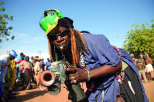 Prozession während der Feierlichkeiten zum Festival sur le Niger: Das Foto zeigt eine dunkelhäutige afrikanische Person mit auffälliger Cappy, braunen Dredlocks und vieler Ketten. Er hat eine Sonnenbrille auf und im Mund eine goldene Trillerpfeife, die mit einer Art selbstgebastelter Trompete in der Hand verbunden ist. Im Hintergrund blauer Himmel und viele andere Menschen auf dem Fest.