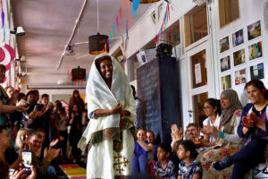 Fashion show in Trampoline House during its ninth birthday in 2019. The photo shows a catwalk improvised in the hallway on which a cheerful dark-skinned woman wears a white traditional robe. The spectators and children are clapping or filming with their smartphones.
