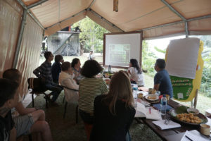 Das Artistic Team der documenta fifteen bei seinem ersten Treffen in Tanakita, Indonesien. Das Foto zeigt eine Gruppe Menschen die an einem Tisch, draußen unter einem Zelt sitzen und auf einen digitalen Projektionsbildschirm schauen. Notizen, Trinken und Essen liegen auf dem Tisch.