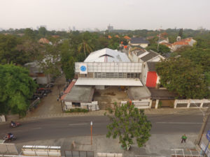 Gudskul in Jakarta, Indonesia. Gudskul in Jakarta, Indonesia. The photo shows from above the building with a blue logo on which Gudskul stands. In front of it a street with passing scooters. The building is located in a built-up area of the city but with a lot of green trees, palm trees and in the background the skyscrapers of Jakarta.