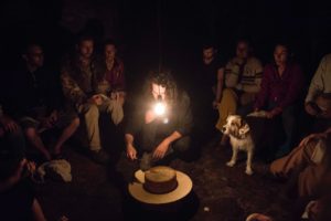 It is dark, only a light bulb hanging down in the middle is shining. People are sitting around it and looking into the center, where a large round cheese is lying under the light bulb. This is cut by the male person squatting in front of it. Among the people, a dog also looks excitedly at the action.