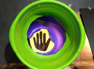 A green ceramic pot from above, on the bottom on purple and yellow a black hand-like symbol.