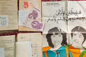 Verschiedene Papierdokumente und Notizen aus den 90er Jahren, darunter eine Zeichnung auf einem kariertem Blatt: zwei Schüler*innen und darüber eine arabische Überschrift.