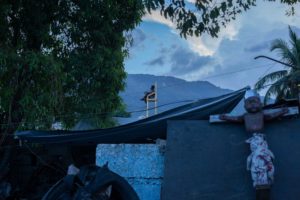 On the roof of a Haitian housing estate, a man sits on a large wooden chair surrounded by trees, forest in the background, and a crucified, suspended doll in the foreground.