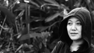 A black and white photo of the artist. She has a hood on and looks into the distance.