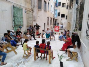 Residents and children sit in a circle in a courtyard and talk to each other. The Tunisian flag hangs on a clothesline above them.