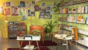A room furnished in 60s style, with green, posterized walls. Next to a large bookshelf, piles of books lie on small tables in the middle. In the back on the wall shelf there's a radio, a typewriter and a telephone.