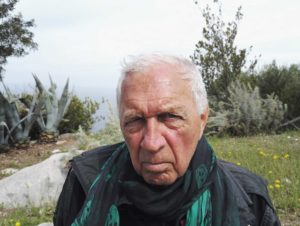 A photo of Jimmie Durham, looking directly into the camera with his right eye squinted shut, wearing a black bandana with green skull heads.