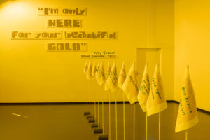 Ein gelb beleuchteter Ausstellungsraum. Auf der Wand steht ein Zitat 