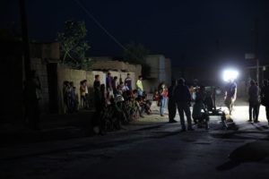 It is night and a film scene is being shot outdoors. Spotlights illuminate the camera team. Children and residents stand or sit along a wall and watch the filming.