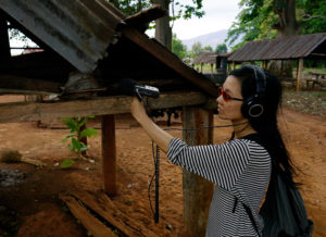 Die Künstlerin, mit Sonnenbrille und Kopfhörer, hält ein Aufnahmegerät in ausgestreckter Hand. Im Hintergrund dörfliche Holzhütten.