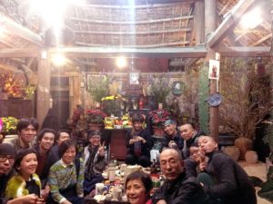 Ein Gruppenfoto während des vietnamesischen Neujahrfests. Die Menschen sitzen in einem Holzhüttenraum auf dem Boden um einen kleinen Tisch und schauen fröhlich in die Kamera. Im Hintergrund viele Blumen, Lichter und Töpfe.