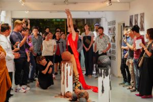 A group of visitors stand in the exhibition space and look at a man standing in the middle. He is wearing a long red vest and black laced high-heeled sandals and is stretching his arms upwards. In the foreground are two round sculptures on two pedestals. Some of the spectators take photos or videos with their cell phones.