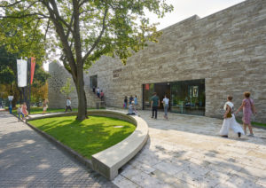 The photo shows an exterior view of the entrance area of the Grimmwelt Kassel, consisting of a white-light brown stone facade. In front of the building a small green area with a tree, sun rays hitting the light stone floor, a few people in front of the entrance.