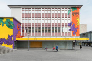 Das ruruHaus in Kassel: Das Gebäude hat viele Fenster, links und rechts an den fensterlosen Flächen ist das Gebäude mit den Kontrastfarben gelb, lila, grün und orange bemalt. Illustriert mit vielen Händen die ineinander greifen.