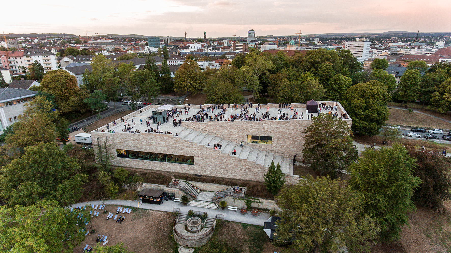 The photo shows the modern building of Grimmwelt Kassel from the bird's eye view. On the roof of the building, which is accessible by wide stairs, there are many people. Around the building many trees and on the horizon the skyline of Kassel.