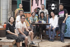 The group photo shows the nine people of the artist collective. They are outside on a kind of terrace on wooden leaves, in the background a container, colorfully painted, in the middle a table with coffee cups and a plant. Some are sitting, some are standing, all are facing the camera. Some look smiling into the camera, some look neutral to serious. All have casual clothes on, mostly a T-shirt and jeans.