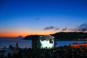 A film screening on a big screen on the beach at night with a lot of viewers.