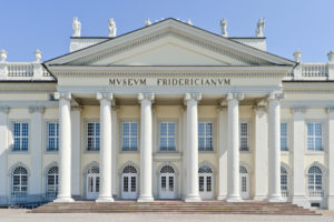 Das Foto zeigt die weiße klassizistische Fassade des Fridericianums, dahinter blauer Himmel. Die Front des Gebäudes ist durch einen von sechs ionischen Säulen getragenen Portikus geprägt. Über dem Eingang steht in großen goldenen Versalien 