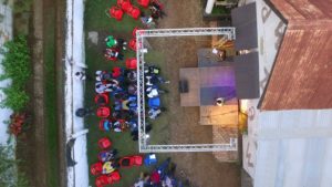 A bird's eye view directly from above of an open air stage next to a building. The stage is framed by a white scaffolding. Two people are standing on the stage. In front of the stage red chairs, some empty, some with visitors.