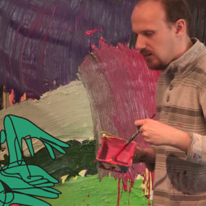 A man holds a brush and container with red paint. In the background his picture with thick green-purple paint layer.