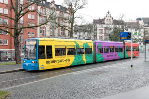 Straßenbahn im documenta fifteen Design. Drei Wagons jeweils gelb, lila, rot; dazwischen die grünen Hände.
