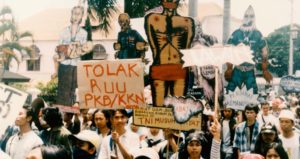 Photo of a demonstration against militarism. People hold up signs and pap figures being tortured.