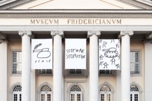 Zwischen den Frontsäulen des Museums hängen drei Banner mit Zeichnungen eines Panzers und der Aufschrift 