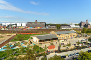 The photo shows the building of the Center for Art and Urbanistics in Berlin from above . It is made out of light brown bricks and has a large courtyard area in the front, also a lot of greenery. In the background some railroad lines, behind them more buildings, blue sky.