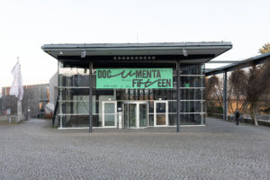 Das Foto zeigt die documenta Halle mit ihrer großen Glasfront, den Stahlträgern und einem Flachdach. Vor der Eingangsdrehtür hängt ein großes grünes Banner mit dem Schriftzug 