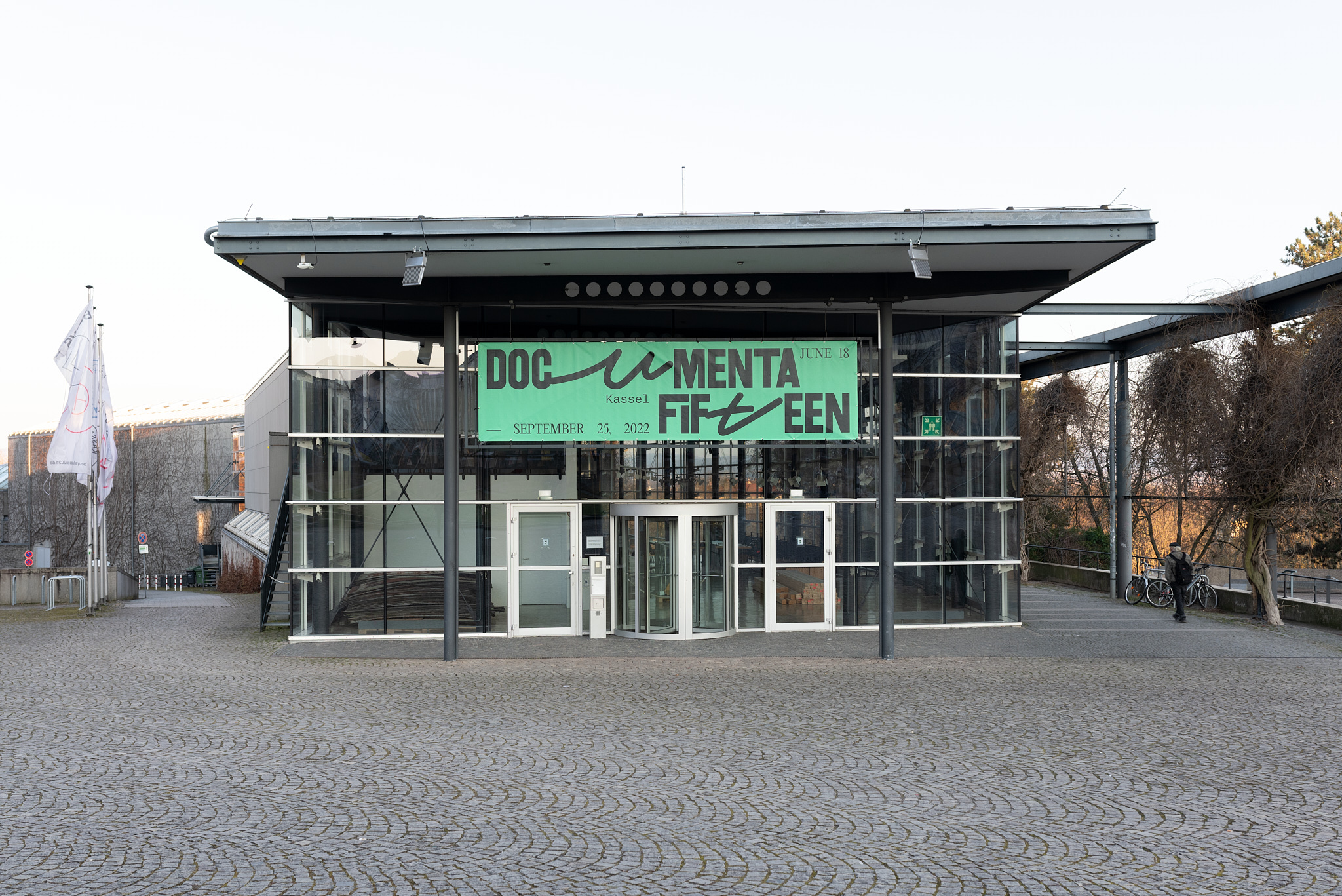 Das Foto zeigt die documenta Halle mit ihrer großen Glasfront, den Stahlträgern und einem Flachdach. Vor der Eingangsdrehtür hängt ein großes grünes Banner mit dem Schriftzug "documenta fifteen".