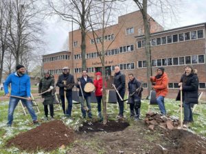 Neun Menschen mit Schaufeln in der Hand stehen symbolisch vor einem frisch gepflanzten Baum.