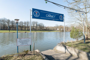 Das Foto zeigt das Ufer der Fulda mit Blick zum Fluss an einem sonnigen Tag. Über einer kleinen betonierten Anlegestelle wird von zwei Stangen ein großes blaues Schild mit dem Schriftzug