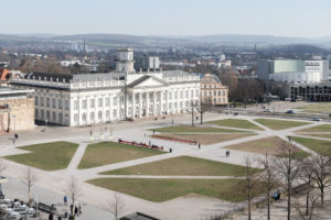 The photo shows Friedrichsplatz and its surroundings from above. You can see green areas, paths, the State Theater, the Fridericianum, trees, a few people and the city, as well as small hills silhouetted on the horizon.