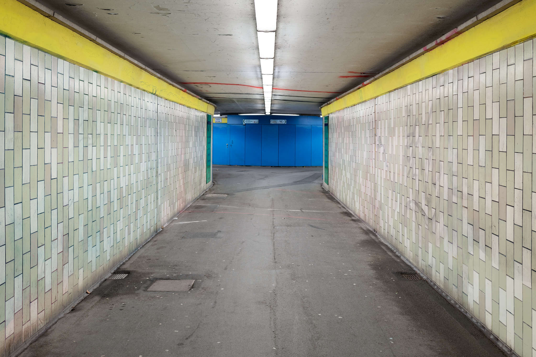The photo shows the tunnel of an underpass. On the right and left a wall with white and green tiles in 50s style, bright neon light at the top of the ceiling, the floor gray concrete, the back wall in bright cobalt blue.