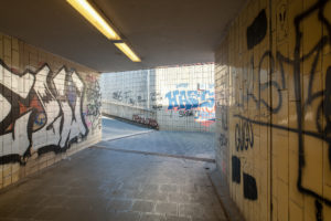 Zu sehen ist eine Fußgängerunterführung. An den bekachelten Wänden sind große Graffitis, oben an der Decke leuchtet eine Neonröhre, Sonnenstrahlen am Ende des Tunnels.