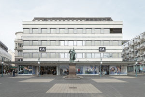 The photo shows the plain and ornamentless facade of the C&A store. In front of it a gray concrete square with an old weather-beaten statue on a pedestal.