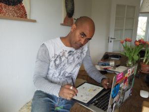 Abdul Dube sitzt in einem Zimmer am Schreibtisch und skizziert auf seinem Tablet, vor Abdul ein aufgeklappter Laptop.