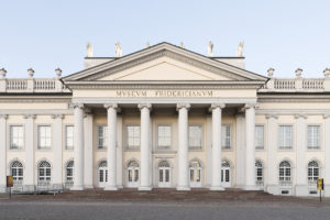Das Foto zeigt die weiße klassizistische Fassade des Fridericianums, dahinter blauer Himmel. Die Front des Gebäudes ist durch einen von sechs ionischen Säulen getragenen Portikus geprägt.