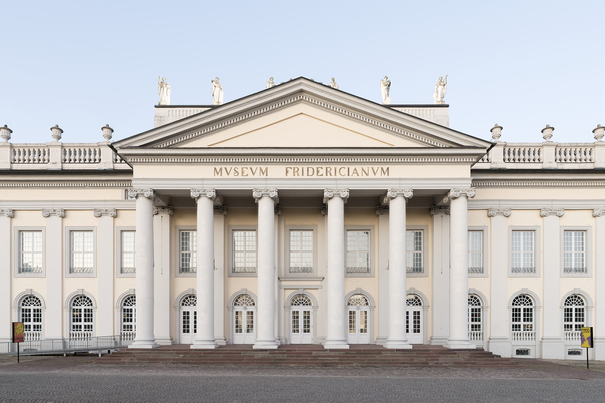 The photo shows the white neoclassical facade of the Fridericianum, with blue sky behind it. The front of the building is characterized by a portico supported by six Ionic columns.