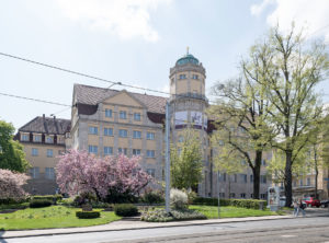 The photo shows the building of the Hessian State Museum. The historicist building has a neoclassical architecture. In front of the building there are small meadow areas and trees.