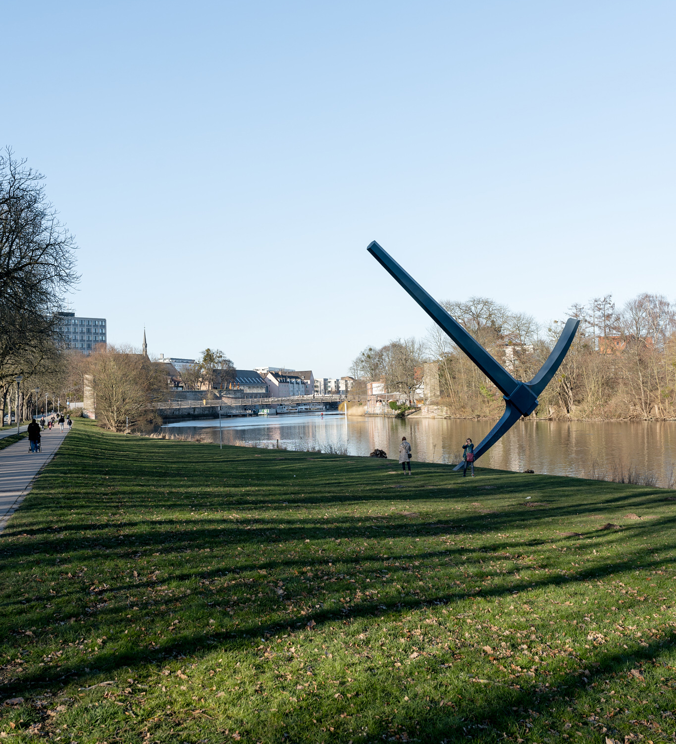 The photo is taken from the bank of the Fulda. It shows green lawn, the blue steel sculpture "Pickaxe" and behind it the Fulda waters, trees, blue sky.