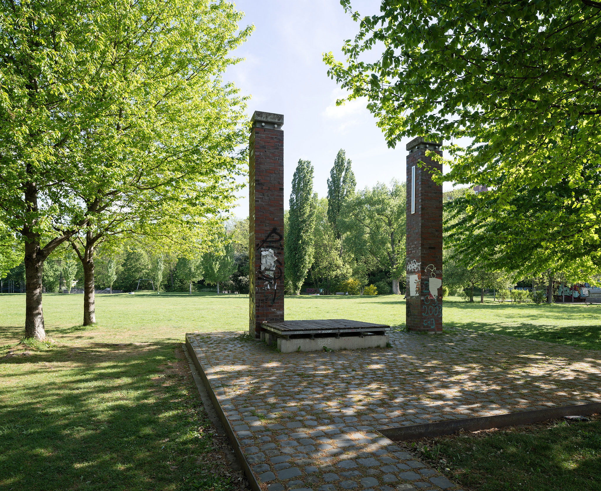 The photo shows the Nordstadtpark. Trees are standing on a green lawn. In the middle you can see a paved square with two pillars made of bricks.